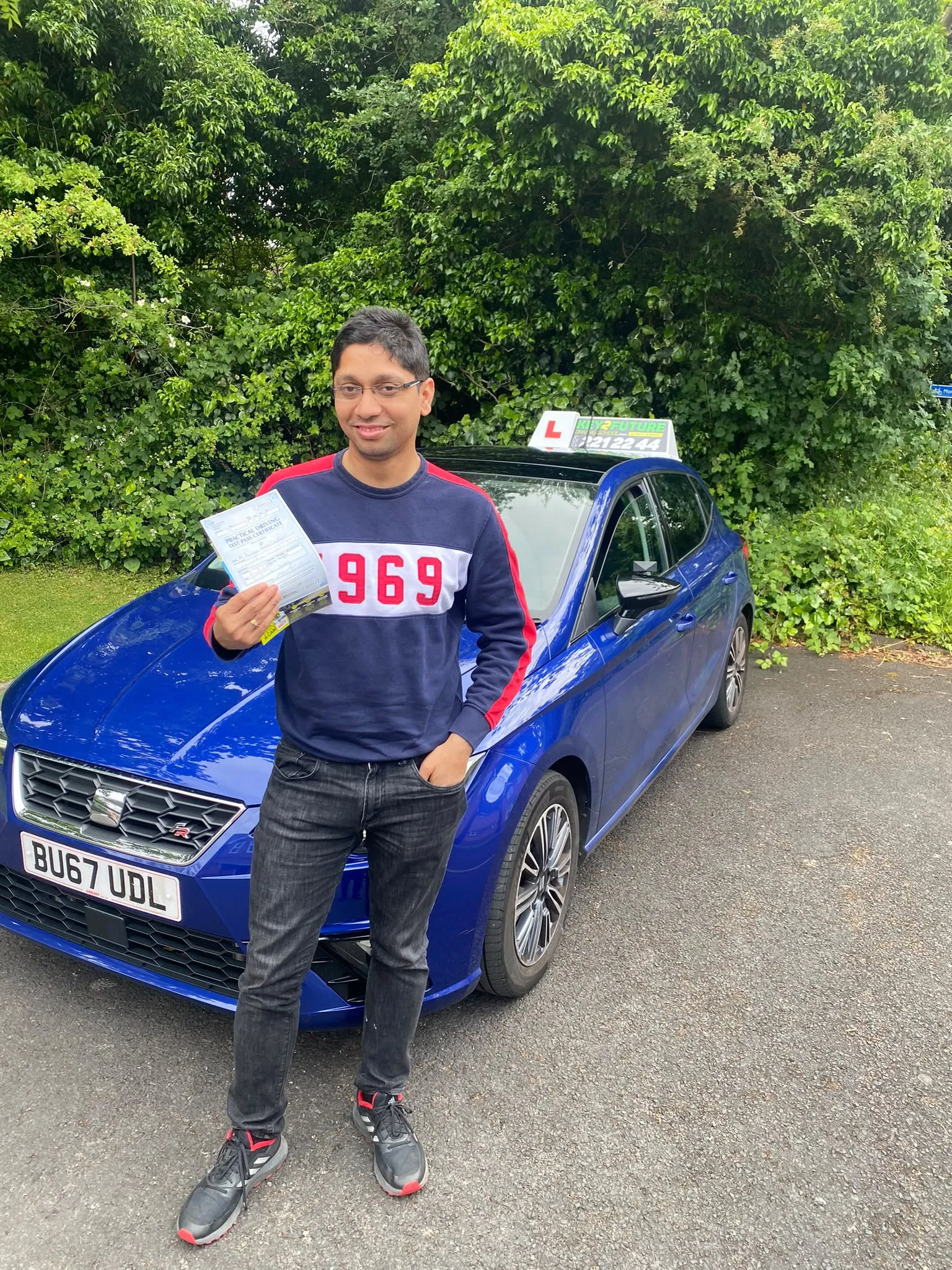 A Key2Future student holding his practical driving test pass certificate beside the school's blue tuition car