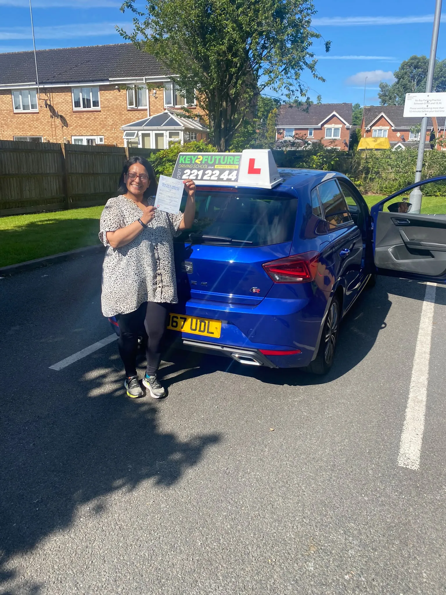 A learner smiling with her driving test pass certificate next to a Key2Future car on a sunny Birmingham street