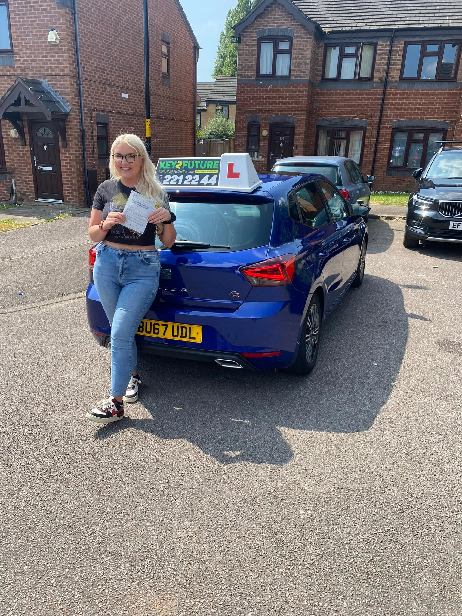 A young woman holding up her practical driving test pass certificate beside a Key2Future tuition car