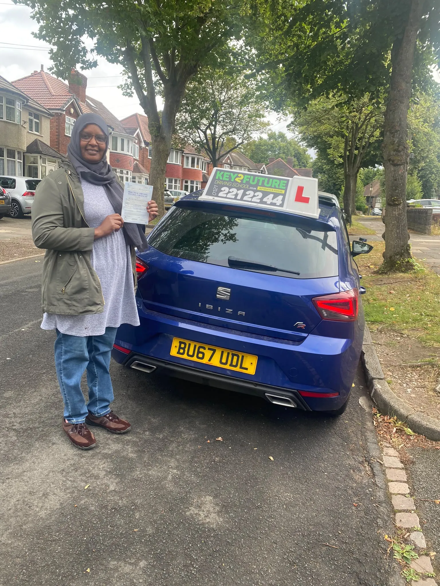 A student holding her pass certificate beside the Key2Future car on a tree-lined Birmingham road