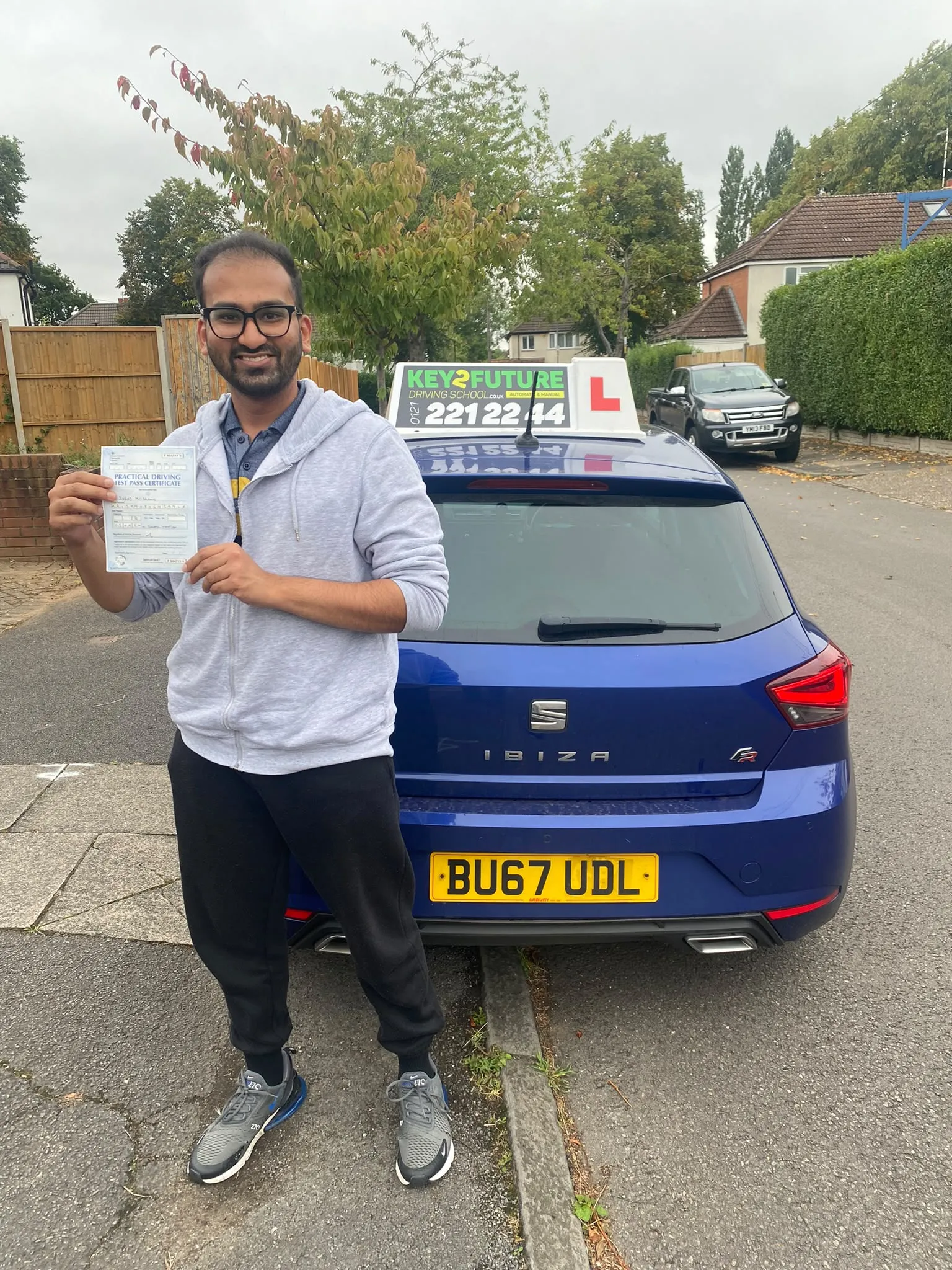 A learner in a grey hoodie holding his practical driving test pass certificate by the school's car